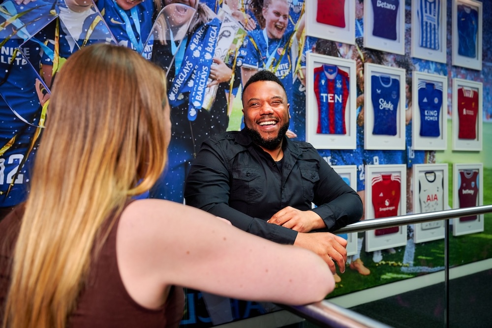 Man in a black shirt smiling and leaning on a table, with football memorabilia and a framed Crystal Palace jersey on the wall behind him.