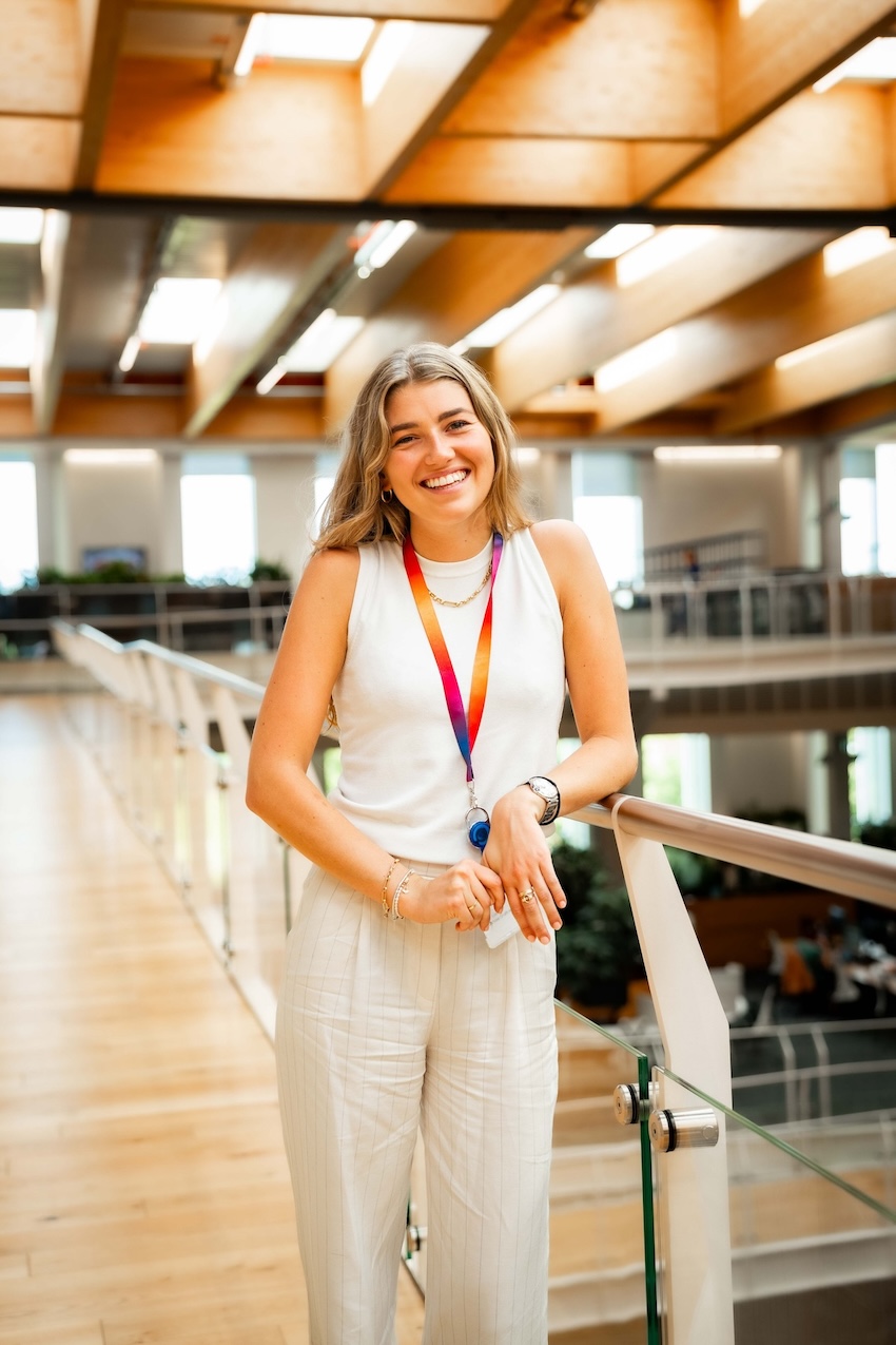 Smiling woman in a white sleeveless top and striped trousers stands indoors by a glass railing, wearing a rainbow lanyard and holding a face mask in a brightly lit modern office.