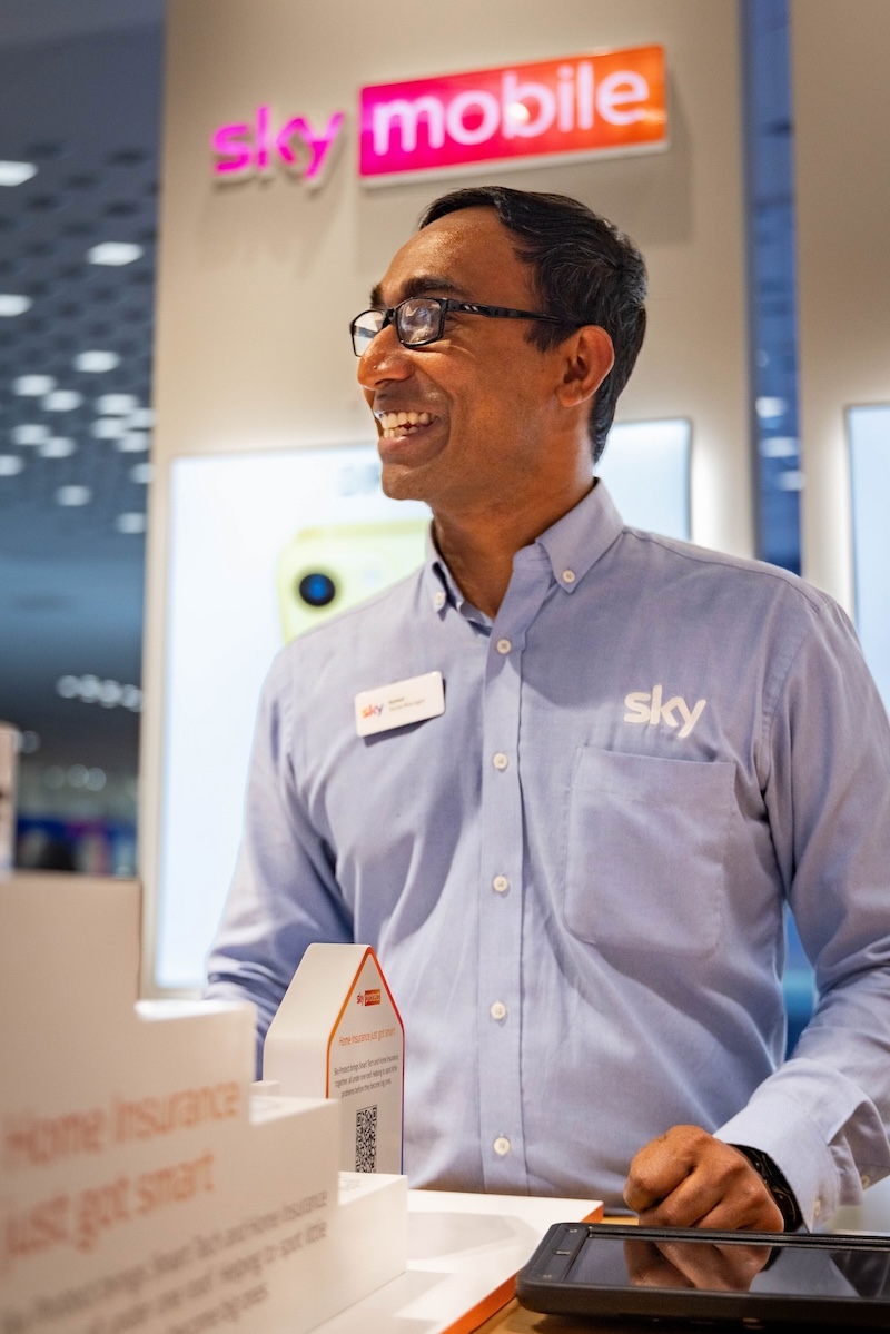 Smiling Sky Mobile employee in a blue shirt stands behind a counter with product displays and a tablet, with bright signage and phone adverts in the background.