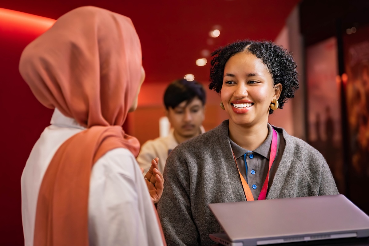 Woman in grey sweater with a laptop smiles while speaking to a colleague in a pink headscarf, with a third person in the background.