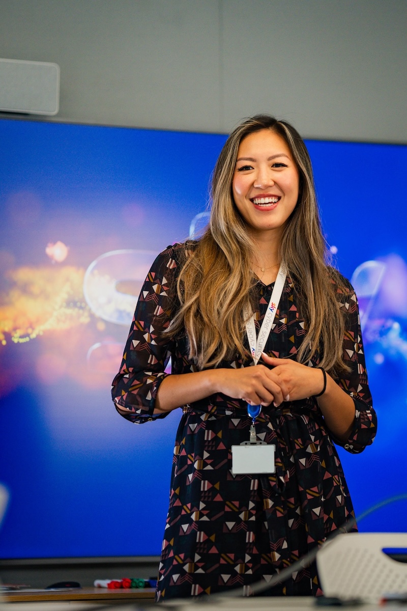 Woman presenting and smiling in front of a blue screen, wearing a patterned dress and a white Sky lanyard.