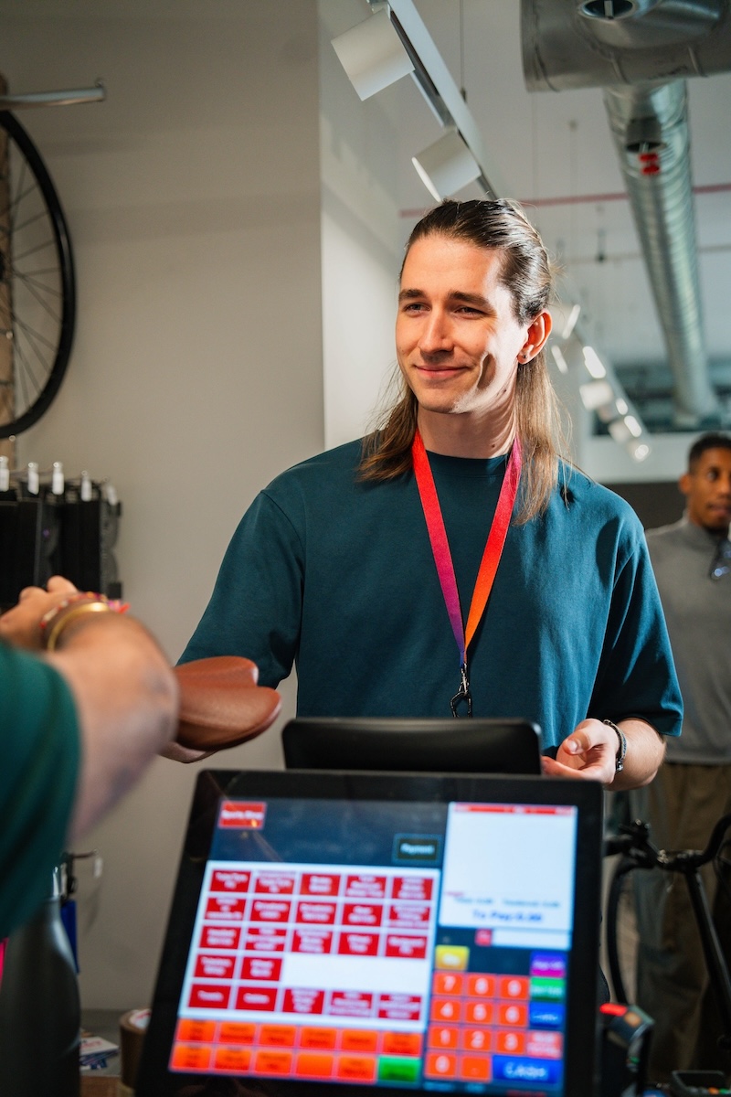 Smiling employee with long hair and rainbow lanyard stands behind a till handing a brown item to a customer in a modern retail space.