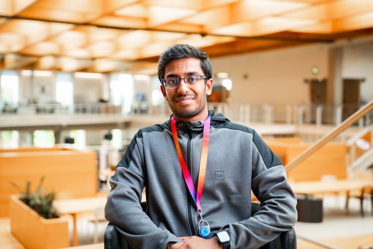 Seated man wearing glasses and a grey hoodie with a rainbow lanyard smiles slightly in a bright, modern office atrium.