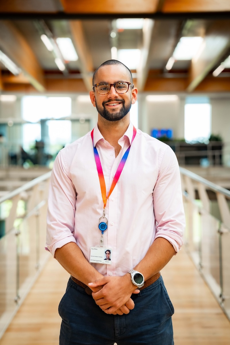 Smiling man in a pink shirt and rainbow lanyard stands in a glass walkway with a wooden ceiling and bright office background.
