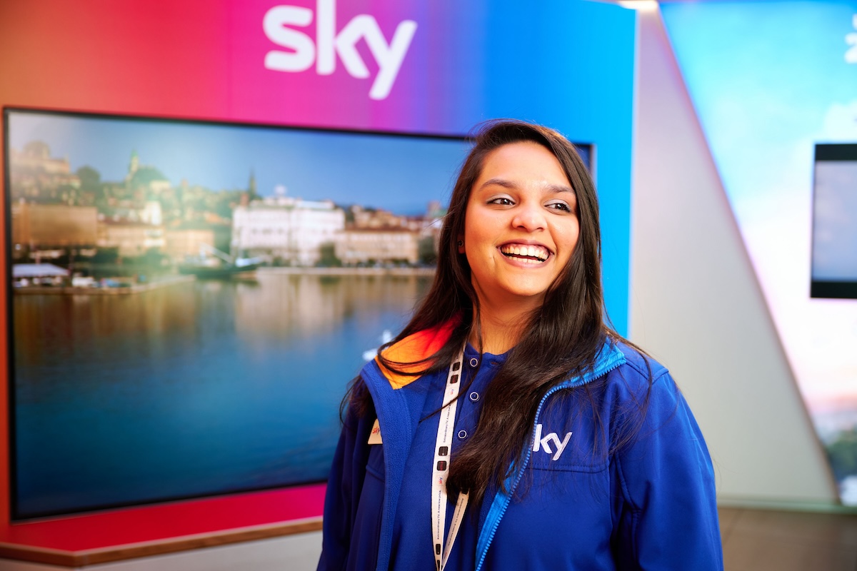 Sky store employee in a blue jacket smiles with a large scenic screen and Sky branding behind her.