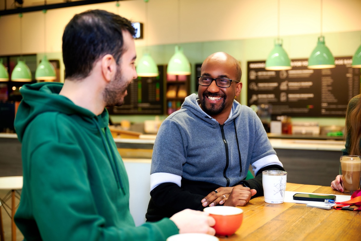 Two smiling men sit at a wooden table with coffee mugs, talking in a café-style setting with green hanging lights and a menu board in the background.