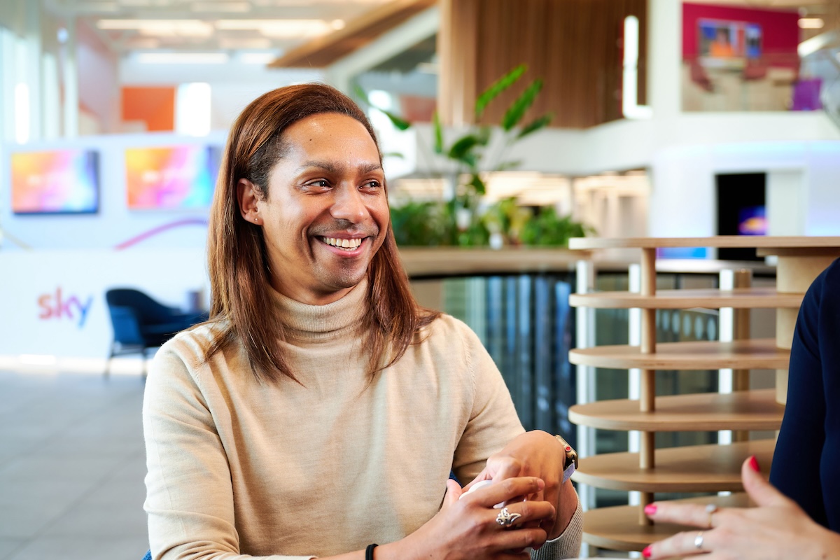 Smiling person in a beige turtleneck sits in a modern, brightly lit Sky office, conversing with someone whose hands are visible in the frame.