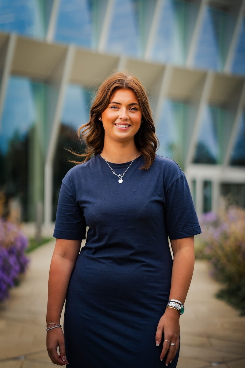 Smiling woman in a navy dress stands on a stone path with purple flowers and a modern glass building in the background.