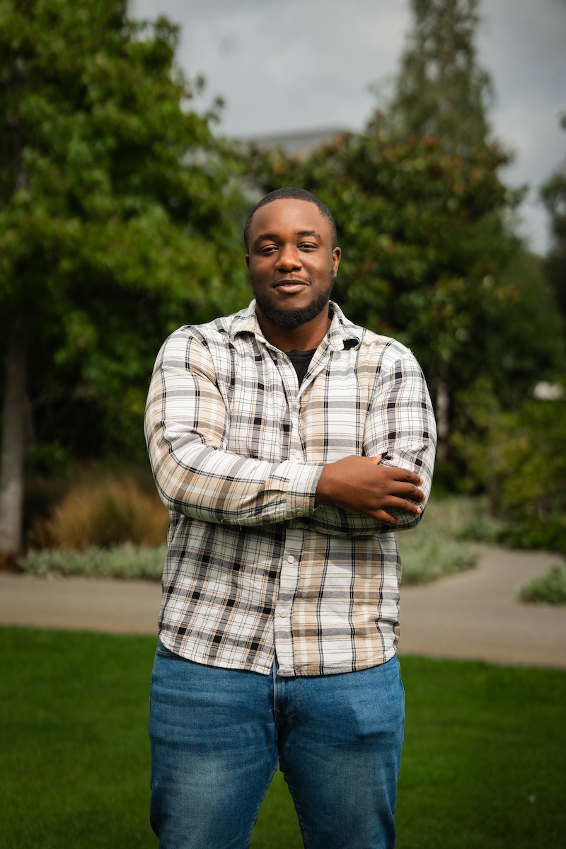 Man in a plaid shirt and jeans stands with arms folded on grass, with greenery and a winding path in the background.