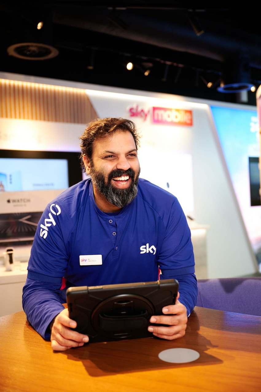 Smiling man in a Sky uniform leans on a table holding a tablet, with a Sky Mobile sign and tech displays in the brightly lit store behind him.