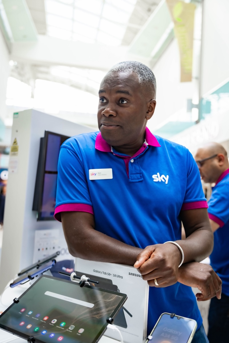Sky employee in a blue and pink uniform stands at a tech display with Galaxy tablets, smiling in a brightly lit shopping centre.