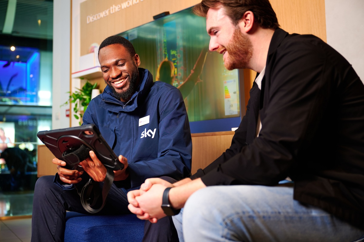 Sky employee in branded jacket smiles while showing a tablet to a seated customer in a retail setting with digital screens behind.