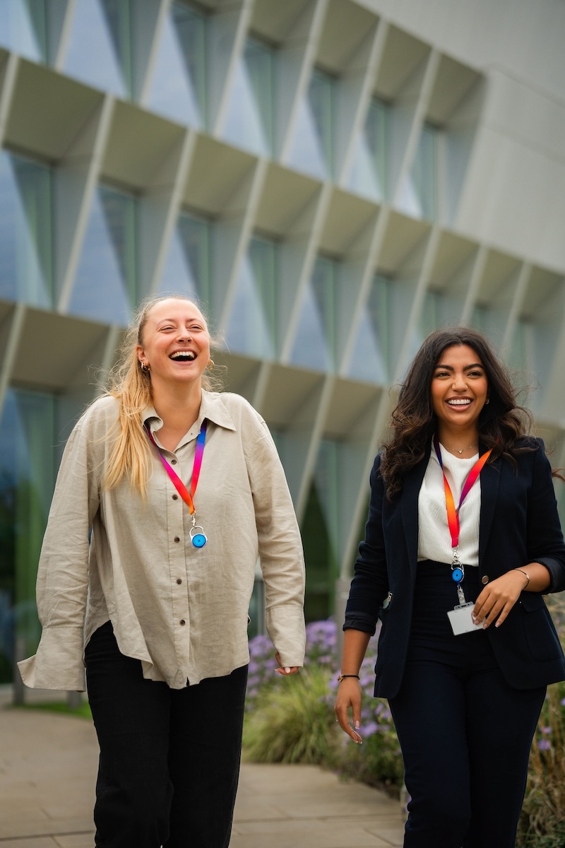Two smiling colleagues wearing rainbow lanyards walk outdoors beside a modern glass-panelled building.