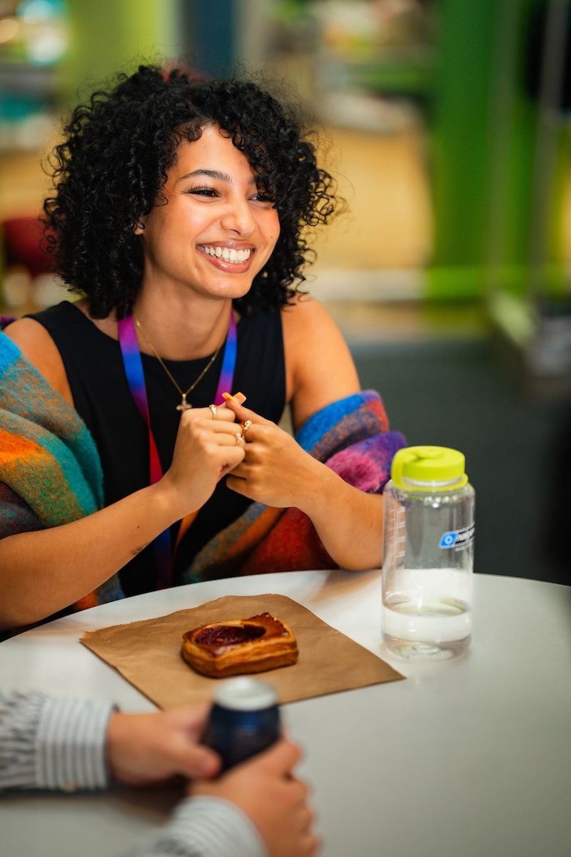 Smiling woman with curly hair sits at a table with a pastry, water bottle, and rainbow lanyard, chatting with someone out of frame.