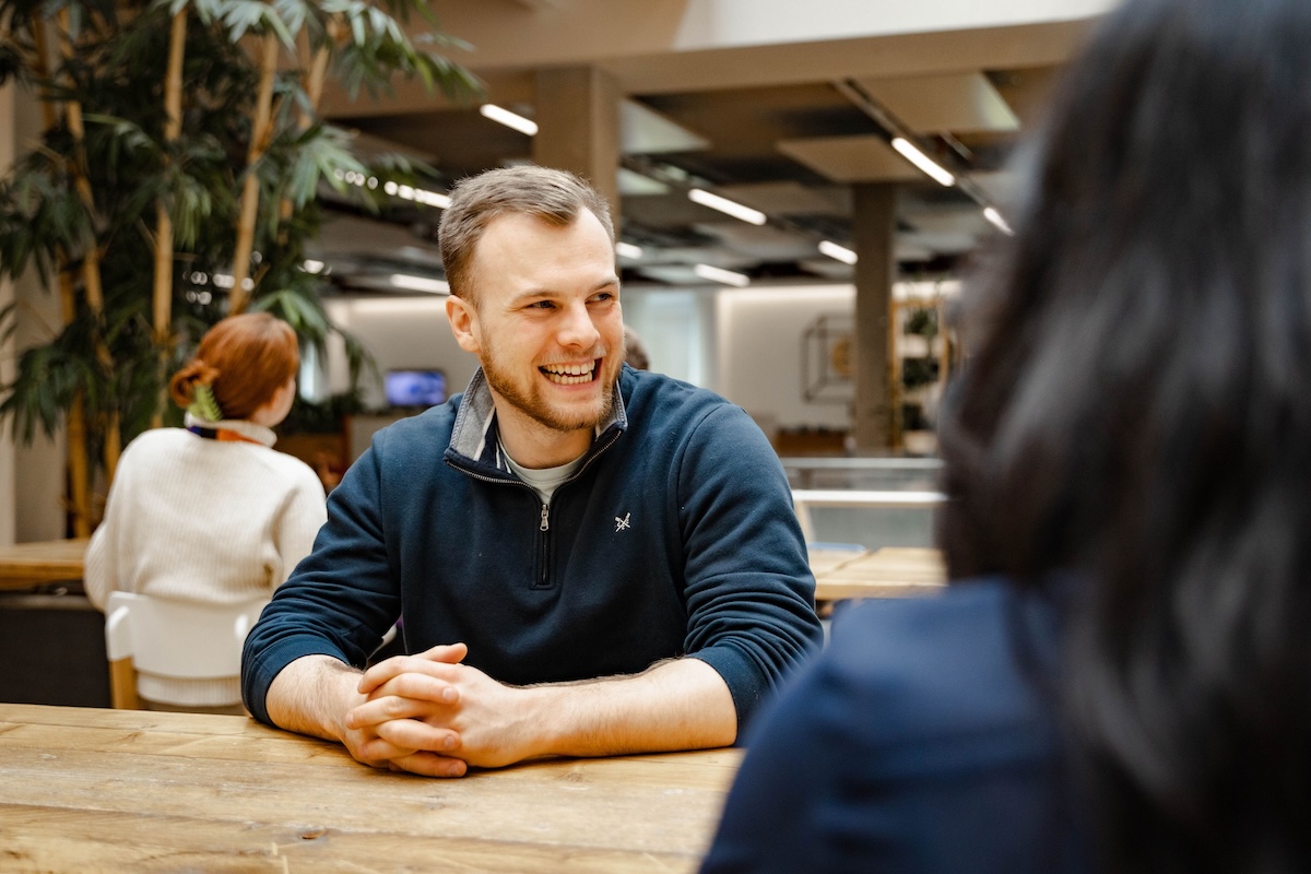 Man with folded hands smiles while chatting at a wooden table in a bright, modern indoor space with plants.