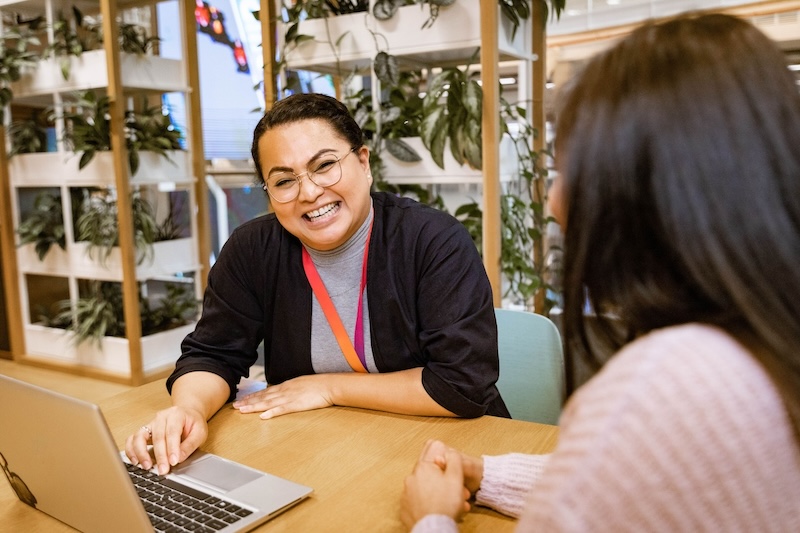 Woman wearing glasses and a rainbow lanyard laughs while working on a laptop across from a colleague.