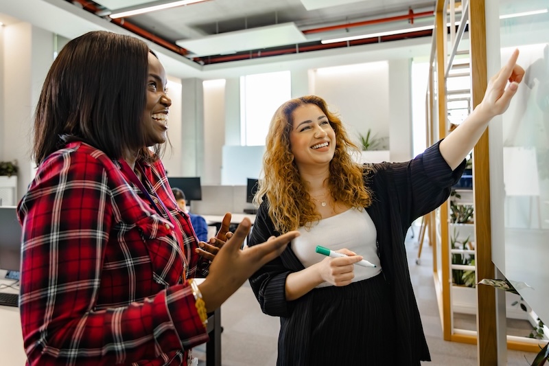 Two women smile while collaborating at a whiteboard in an open-plan office, one pointing as the other gestures.