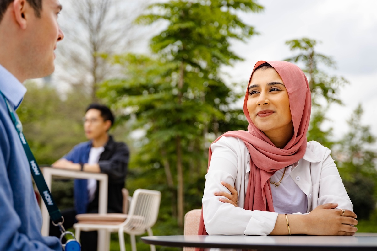 Woman in a pink hijab smiles warmly while talking outdoors at a table with a man, with trees in the background.