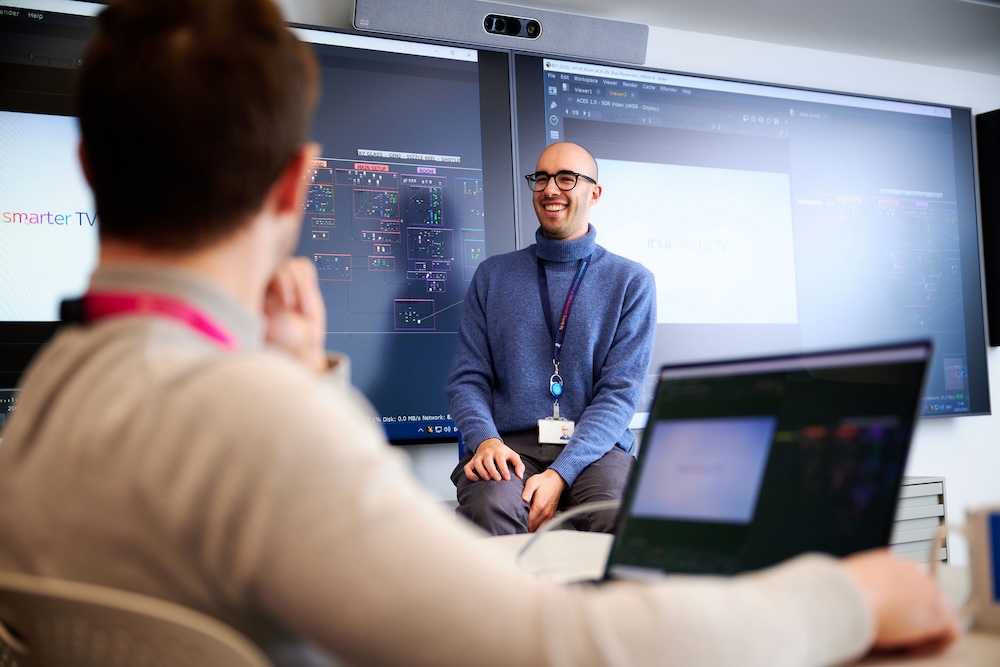 Man with a lanyard sits in front of large screens, smiling while giving a talk to colleagues in a modern tech workspace.