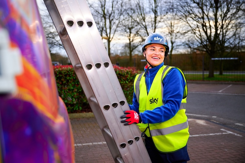 Smiling women in helmet and hi-vis vest stands beside a ladder near a branded van in a car park at dusk.