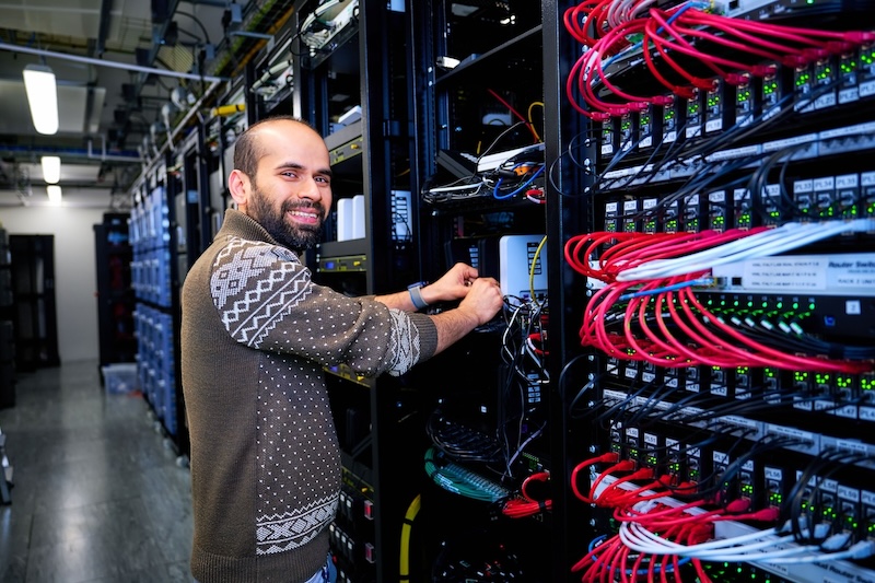 Man smiling while working on a network server rack in a data centre filled with cables and blinking lights.