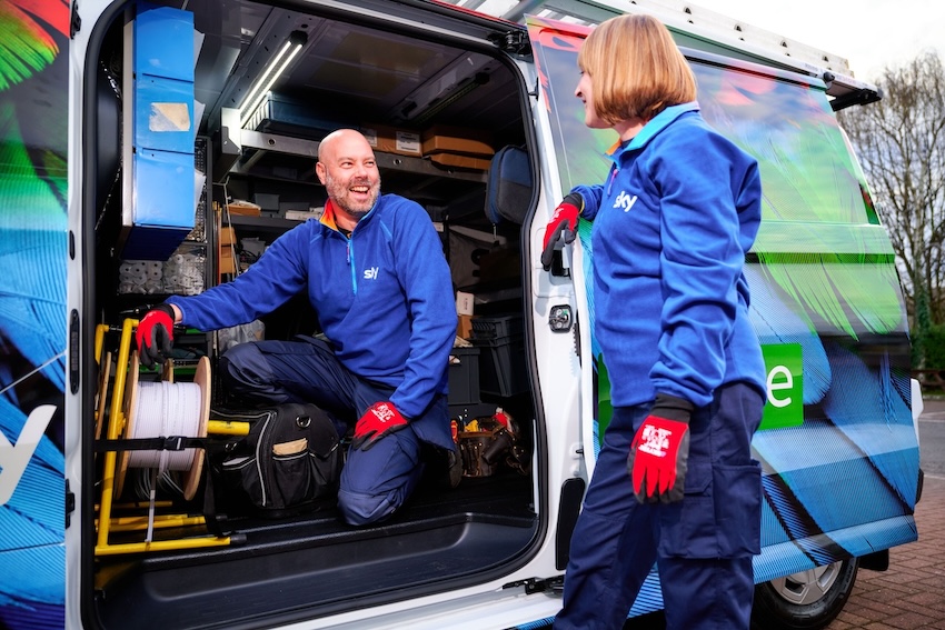 Two Sky technicians in blue uniforms and red gloves talk beside an open service van, with one sitting inside near cable reels and tools, and the other standing outside smiling.