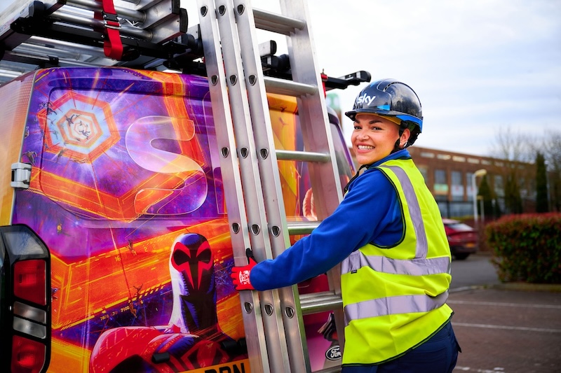 Technician in a helmet and hi-vis vest holds a ladder at the rear of a van decorated with Spider-Man: Across the Spider-Verse artwork, smiling at the camera in a car park.