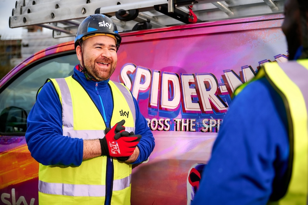 Smiling technician in a helmet and hi-vis vest stands by a van with a Spider-Man design, talking to a colleague out of focus in the foreground.