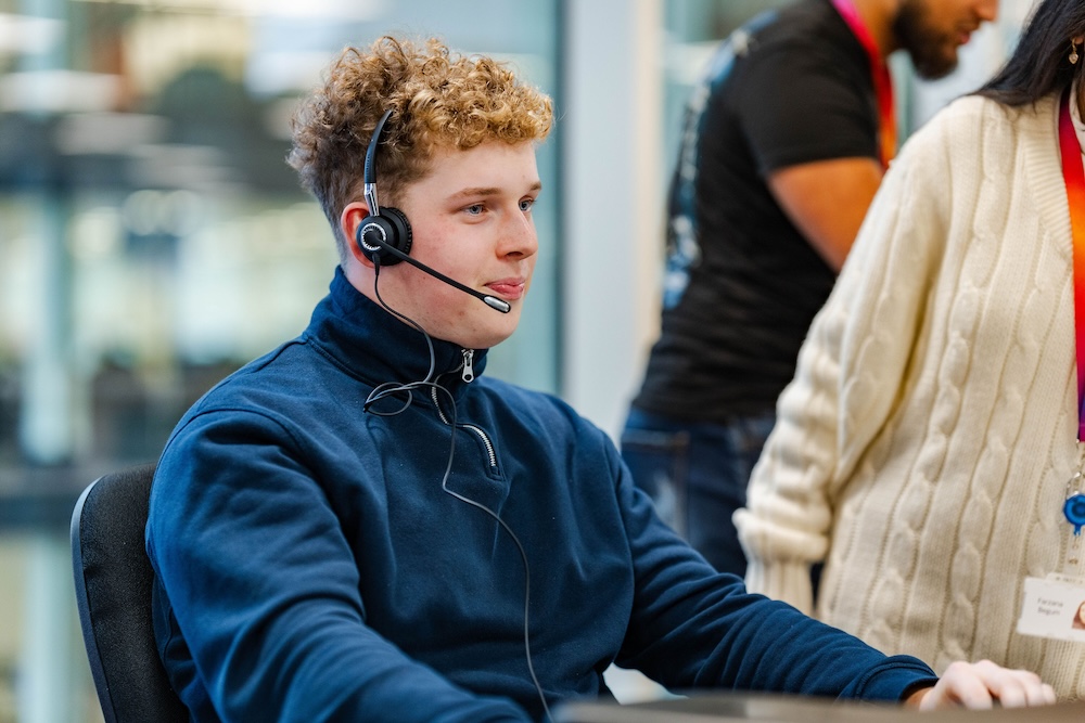 Young man with curly hair wears a headset and navy zip-up in a modern office, seated and engaged, with colleagues in the background wearing lanyards.