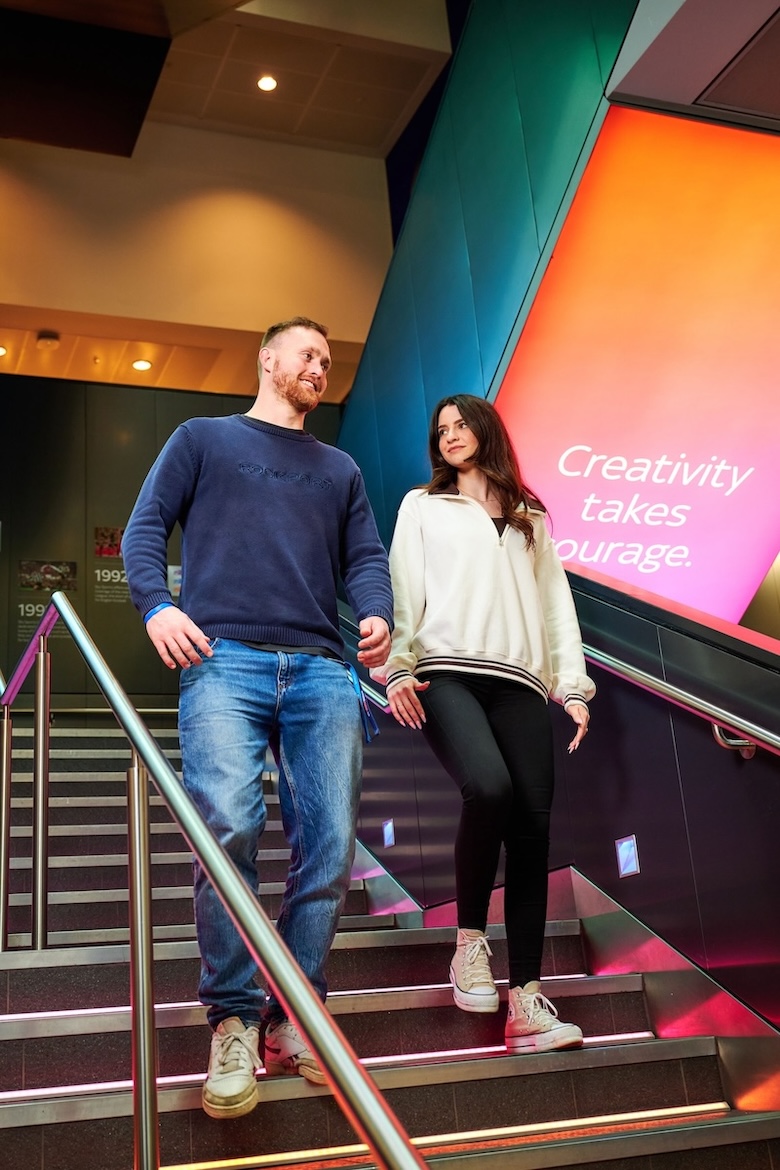 Two people walk down illuminated stairs inside a modern building, smiling at each other, with a bright wall display behind them reading “Creativity takes courage.”