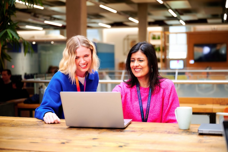 Two women sit at a wooden table in an office, smiling while looking at a laptop screen; one wears a blue top, the other pink, both with lanyards. A mug sits nearby.