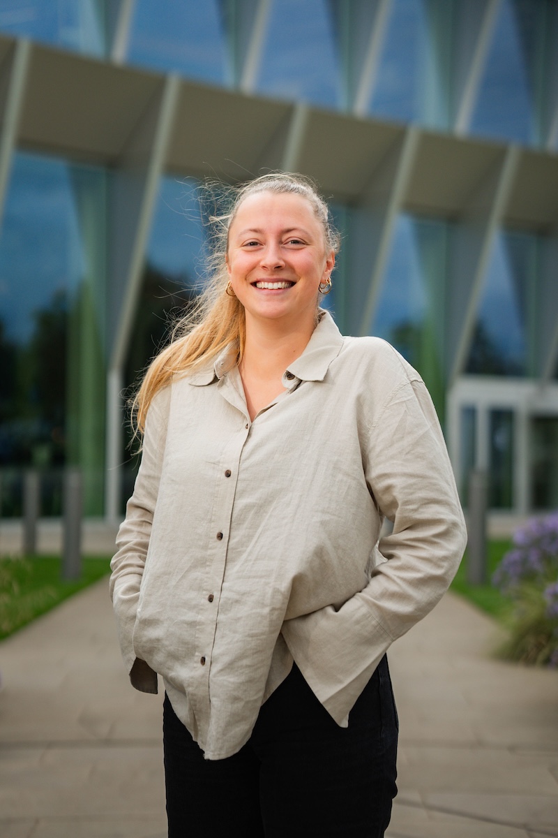 Smiling woman with a ponytail stands outdoors in front of a modern glass building, wearing a beige button-up shirt and black trousers with hands in her pockets.