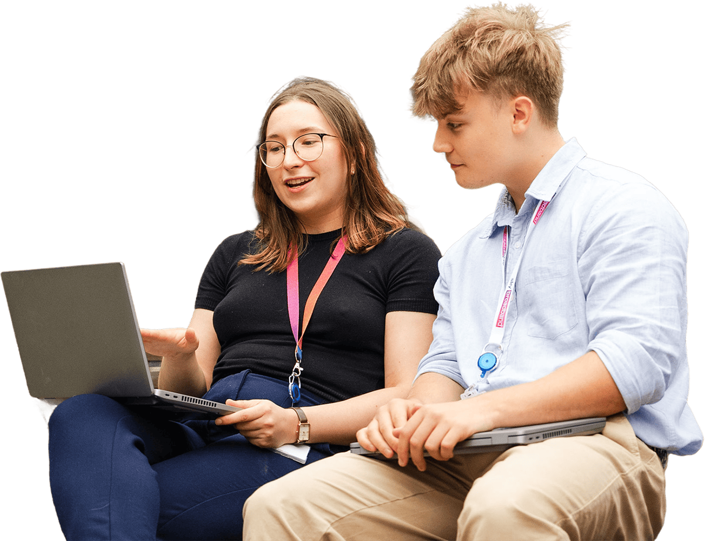 Two young professionals sitting together, collaborating with laptops. The woman on the left is smiling and gesturing at her screen while explaining something, and the man on the right listens attentively with his laptop resting on his lap. Both are wearing lanyards.