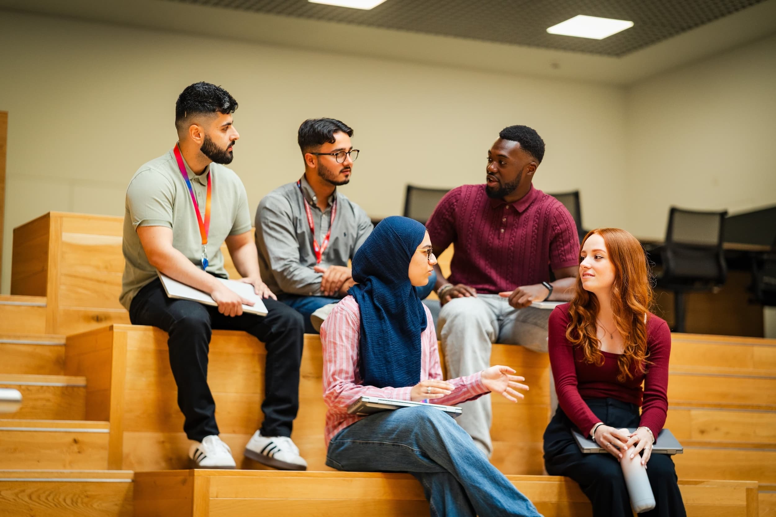 Five young professionals sit on wooden tiered seating, engaged in discussion, each holding laptops or notebooks.