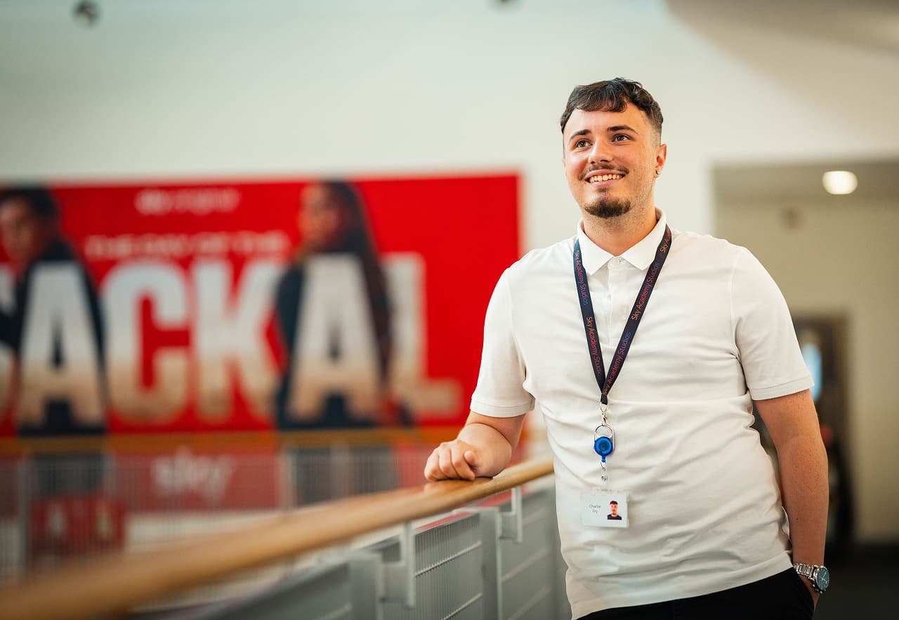 Smiling man in a white polo shirt and lanyard leans on a railing, with a blurred red promotional poster in the background.