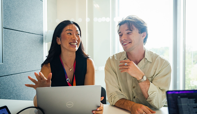 Two colleagues smiling and chatting at a desk with laptops in a brightly lit office near a large window.