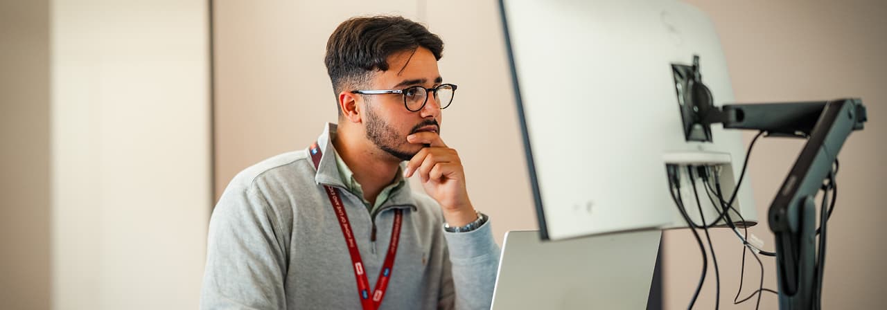 Man wearing glasses and a lanyard working at a desk with two monitors in a bright office.