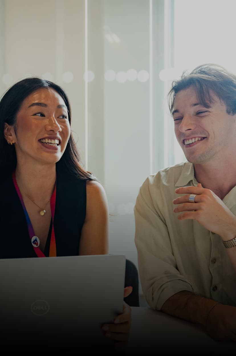 Woman and man smiling while working on a laptop together in a brightly lit meeting room.