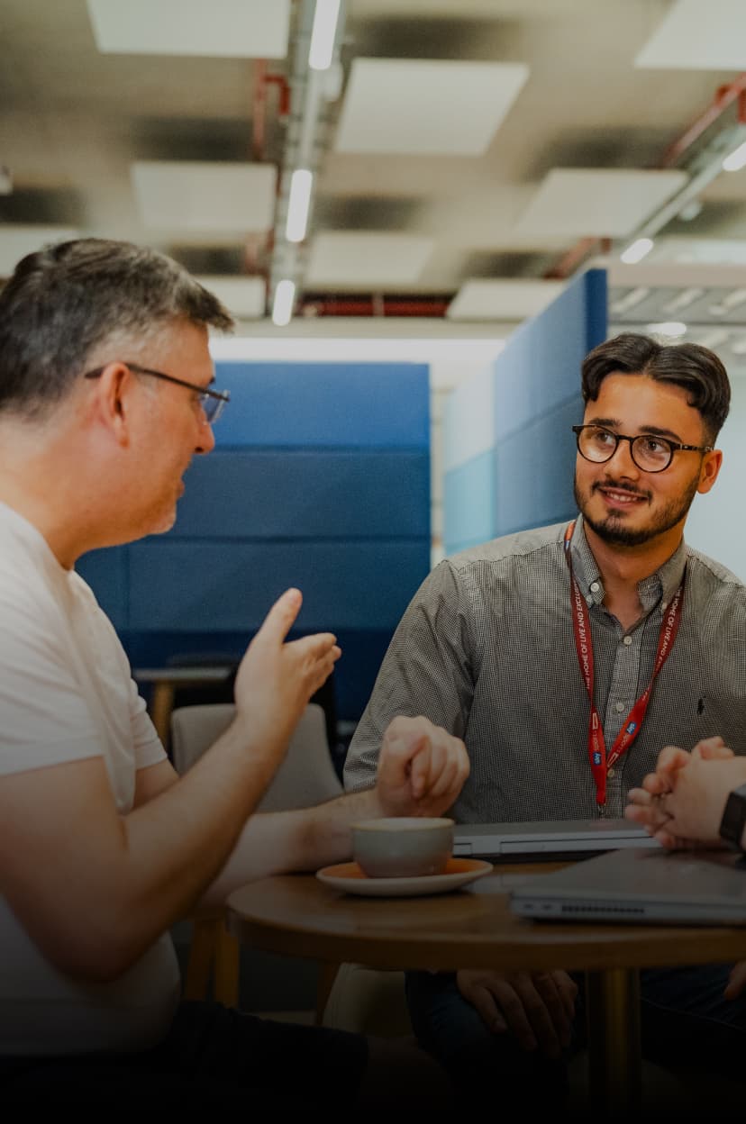 Two men talking at a round table in a casual workspace with coffee cups and a laptop.