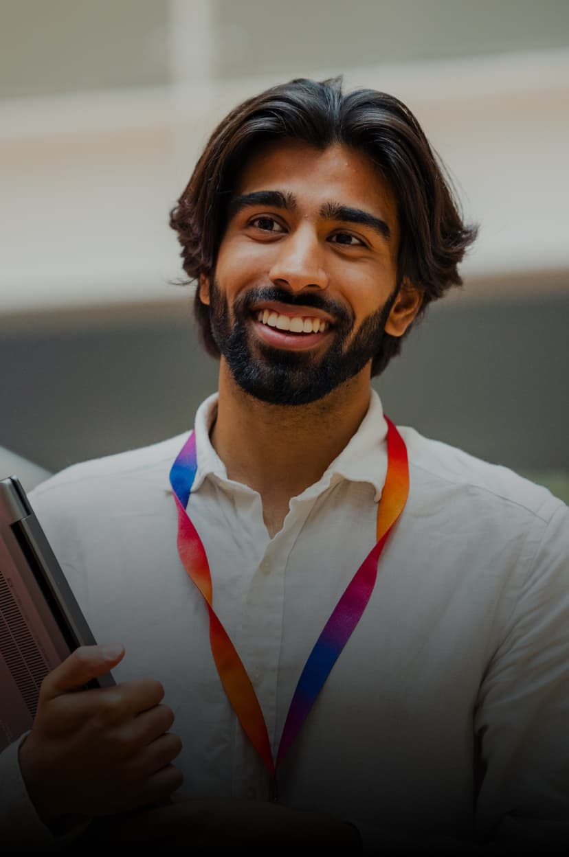 Smiling man holding a laptop, wearing a white shirt and rainbow lanyard, standing in an open office space.