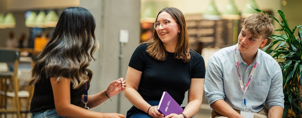 Three young professionals chatting on a bench; one holds a purple notebook, another a reusable cup.