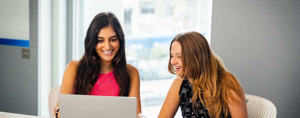 Two women smiling while working on a laptop together at a white desk in a modern office.