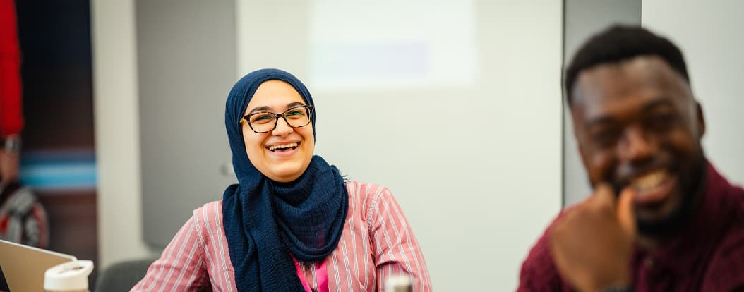 Woman in hijab smiling during a meeting with laptops, seated next to a man who is laughing.
