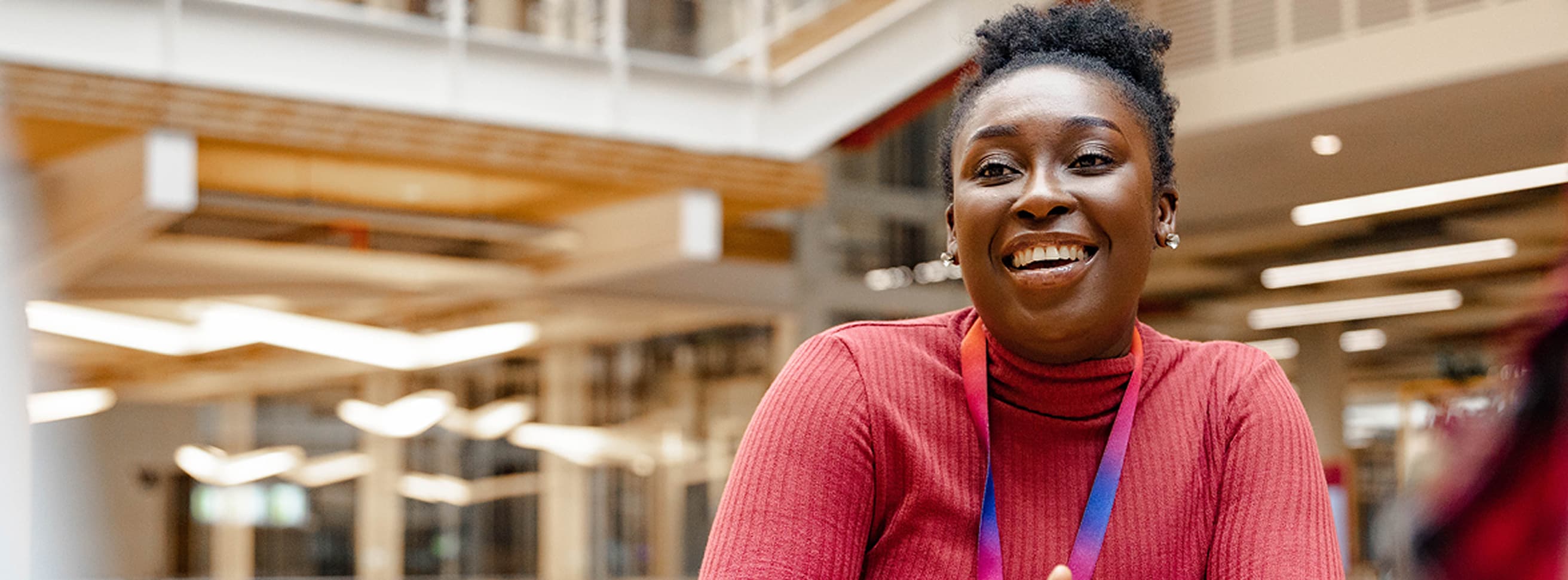 Woman in a red top smiling and leaning forward at a table in a brightly lit modern office space.