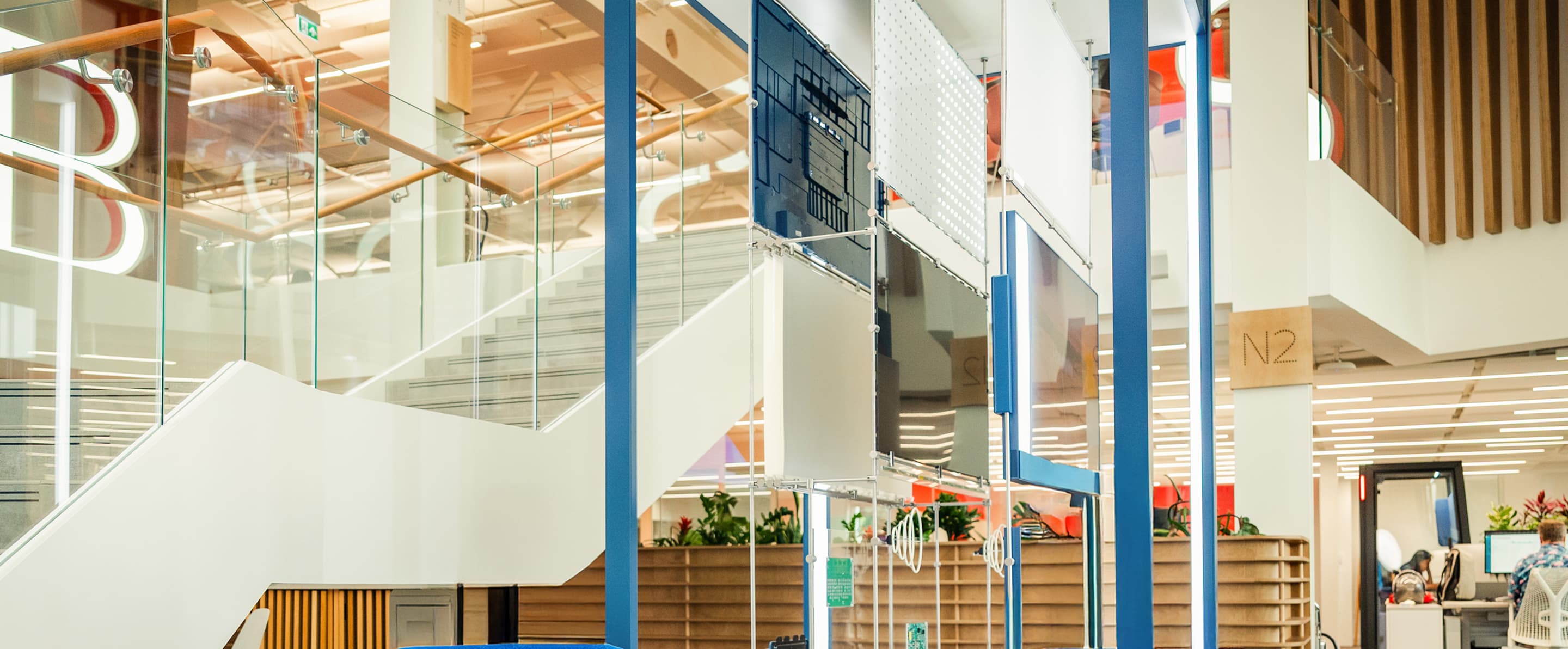 Modern office atrium with glass-railed staircases, hanging panels, and open-plan seating with plants.