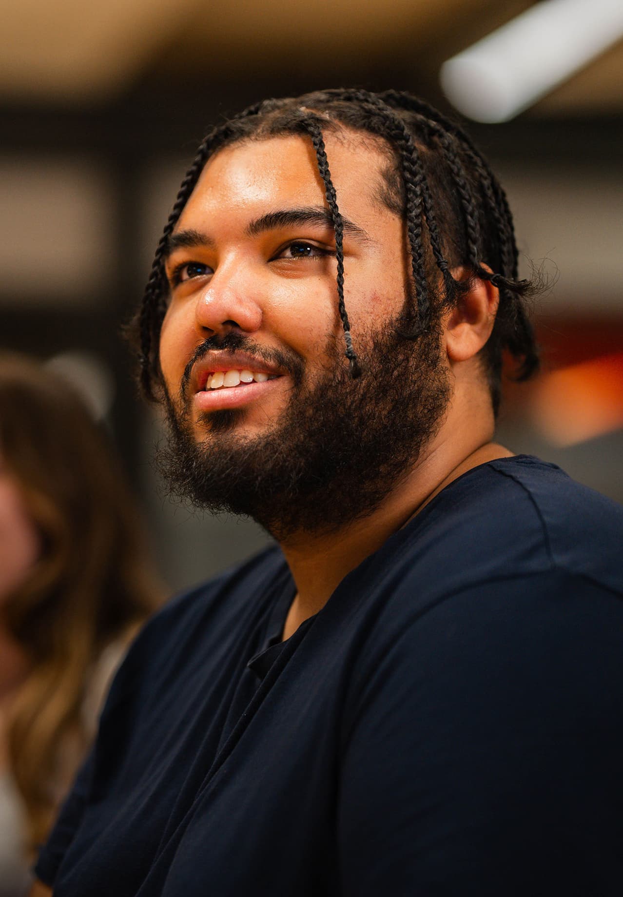 Man with braids and a beard smiling and looking engaged in a casual indoor setting.