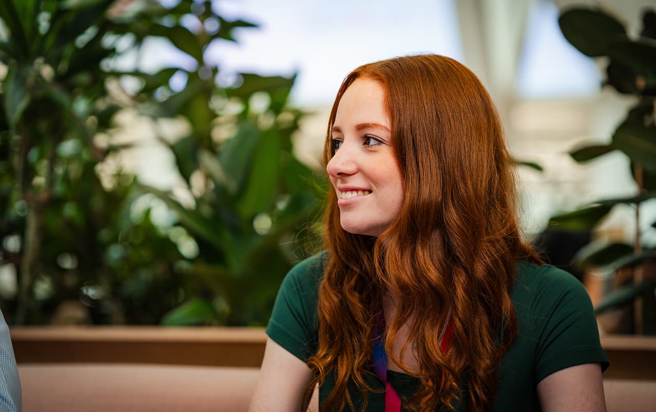 Woman with long red hair smiling in a relaxed setting with leafy green plants in the background.