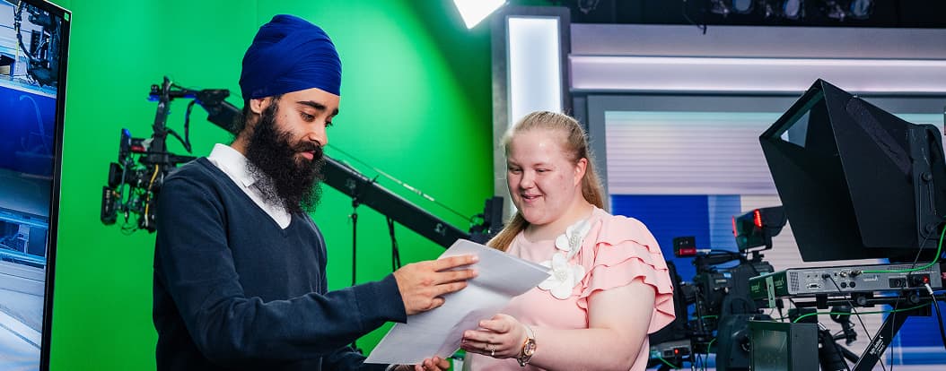 Man and woman reviewing papers together in a professional TV studio with cameras and green screen.