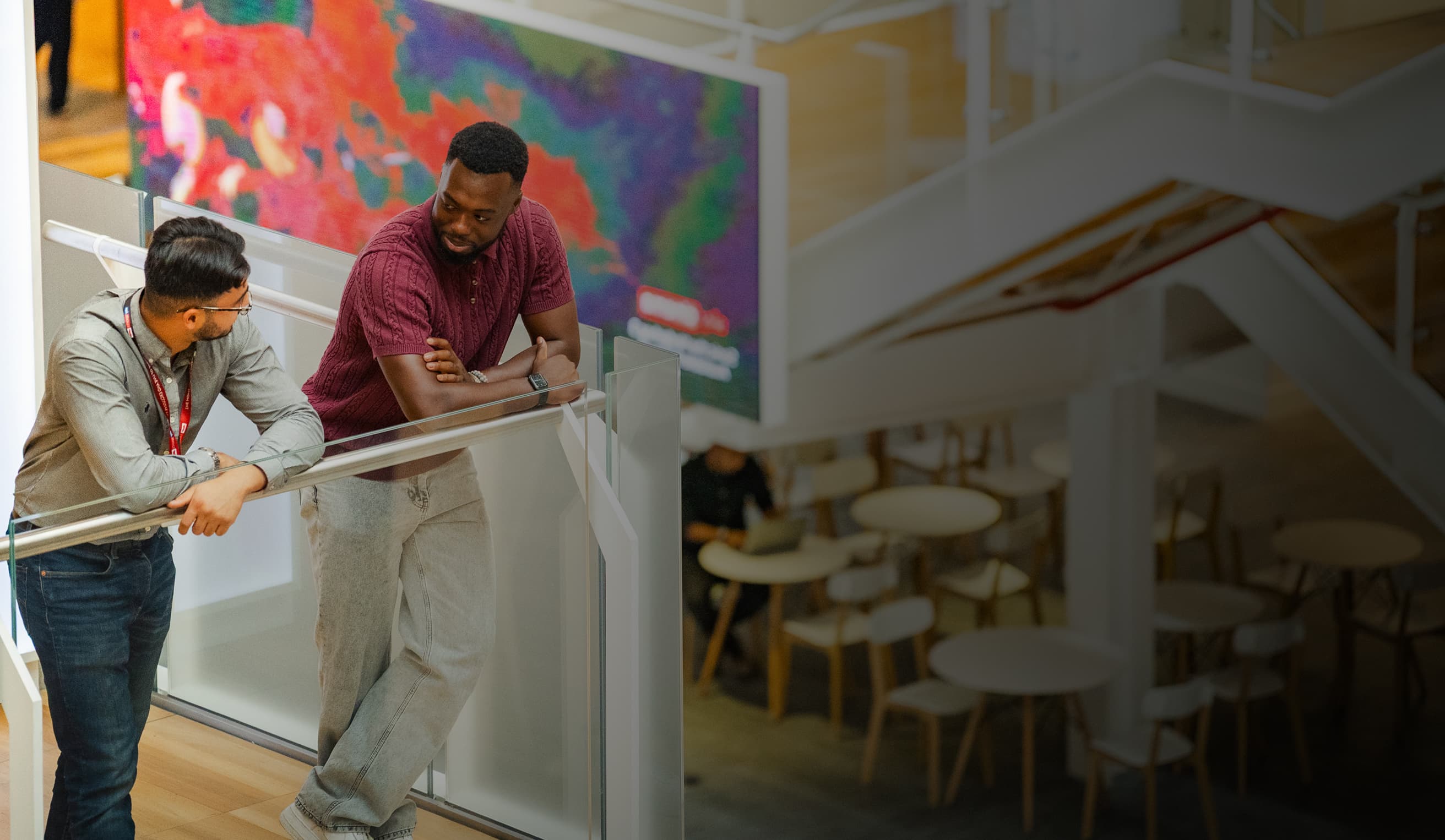 Two men leaning on a railing chatting in a modern office with a colourful digital display behind them.