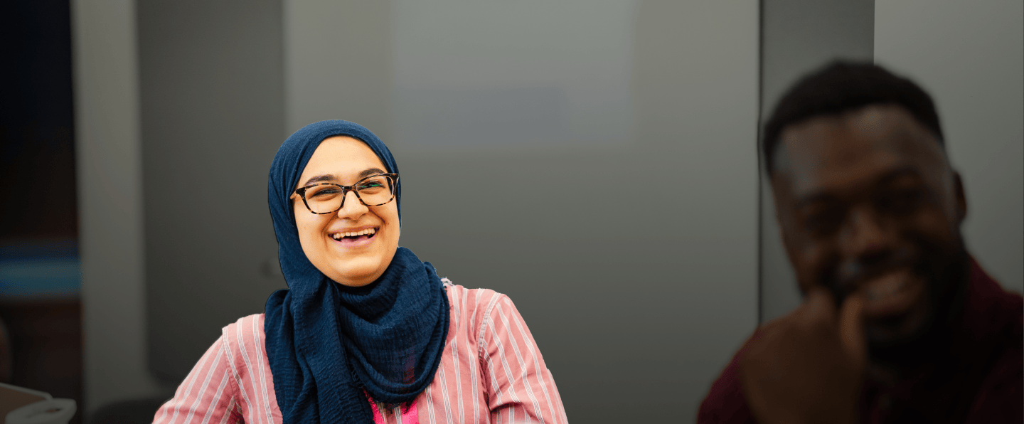 Woman smiling in a meeting room with grey walls, while a man in the foreground appears blurred and smiling.